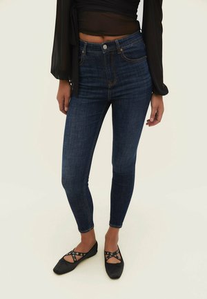 Person iført mørkeblå skinny jeans, sort langærmet gennemsigtig top og sorte flade sko med krydsede stropper og sølvøjne.