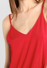 Femme portant un haut rouge sans manches à décolleté en V avec de fines bretelles, montrant une partie de son cou, de ses épaules et de sa poitrine supérieure.