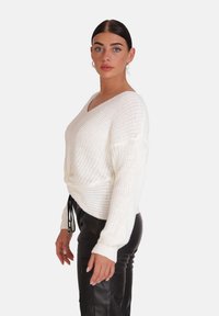 Weißer gestrickter Pullover mit V-Ausschnitt, lockerer Passform und langen Ärmeln. Verfügt über einen Kordelzugsaum und ein strukturiertes Design. Kombiniert mit schwarzen Hosen.