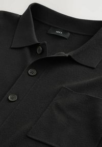Chemise noire à col avec trois boutons noirs et une poche poitrine, avec une étiquette de marque en tissu "NEXT" à l'intérieur du col.