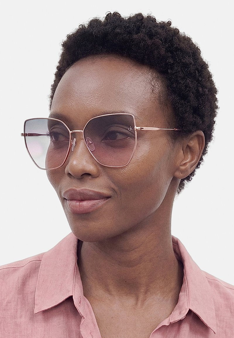 Mujer con cabello corto y rizado que lleva gafas de sol cuadradas grandes rosas y una camisa rosa con cuello, mirando ligeramente hacia un lado.