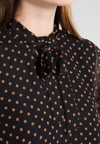 Close-up of black blouse with a tied neck bow and small brown polka dots, showing part of the collar and shoulder.
