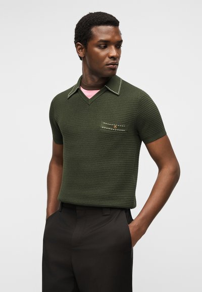 Άνδρας που φορά υφασμένο πράσινο χακί κοντομάνικο πόλο με τσέπη στο στήθος και μαύρο παντελόνι, χέρια στις τσέπες, κοιτάζει προς τα πλάγια.