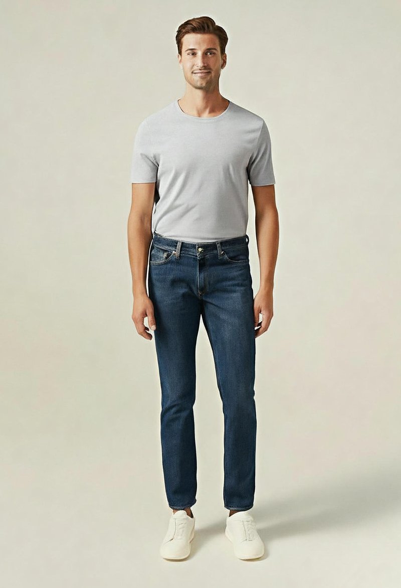 Ein Mann steht da und trägt ein hellgraues T-Shirt, dunkle blaue Jeans und weiße Sneakers vor einem einfarbigen beigen Hintergrund.