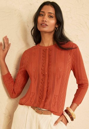 Kvinde med langt mørkt hår iført rust orange knapstrikket top, beige bukser og flere armbånd, stående mod en struktureret beige væg.