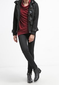 Veste noire à capuche avec boutons à bascule sur un haut rouge à col large, associée à un pantalon noir ajusté et des bottines.