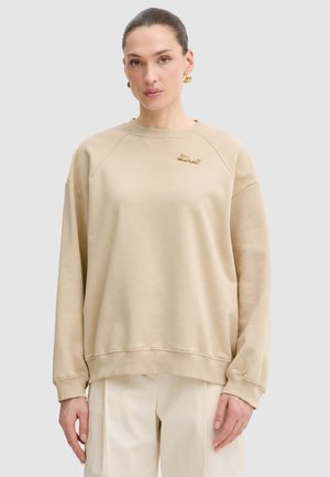 Donna con felpa beige oversize con spilla dorata sul petto e pantaloni bianchi, in piedi davanti a uno sfondo chiaro e uniforme.