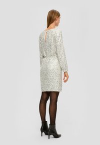 Robe en sequins argentés avec des manches longues, longueur mi-cuisse, taille cintrée, ouverture en forme de clé dans le dos, accompagnée de bottines noires.