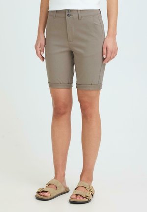 Personne portant un short beige à ourlet roulé mi-longueur au-dessus des genoux et des sandales beige à double bride avec boucles dorées sur fond blanc.