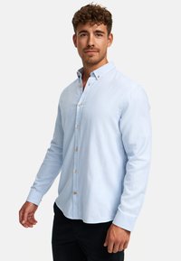 Uomo con capelli ricci che indossa una camicia azzurra a bottoni e pantaloni neri, in piedi con un'espressione neutra su sfondo bianco.