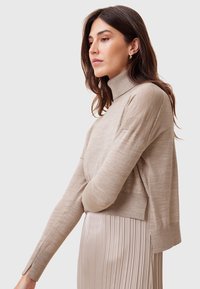 Maglione beige aderente a collo alto realizzato in un materiale morbido, con polsini a coste e spacchi laterali, indossato sopra una gonna plissettata color crema.