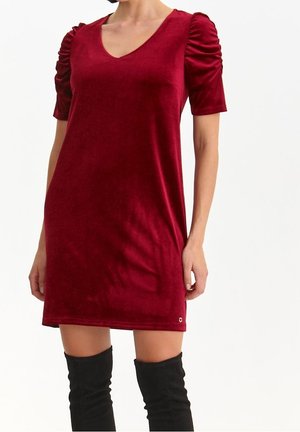 Robe courte en velours rouge à manches courtes avec épaules froncées, associée à des bottes noires montantes, portée par une personne debout.