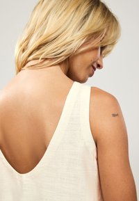 Haut sans manches beige clair avec un dos en V profond, doté d'un tissu texturé et de détails de couture visibles. Petit tatouage sur l'épaule.