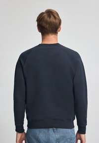 Felpa blu navy con scollatura tonda, maniche raglan e polsini a coste, caratterizzata da una trama morbida e una vestibilità ampia, indossata su pantaloncini di denim.