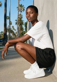 Jong persoon in een wit T-shirt, zwarte korte broek en witte sneakers hurkt op het trottoir naast een muur met palmbomen op de achtergrond.