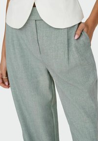 Femme portant un pantalon gris clair cintré avec des plis, la main dans la poche, et un blazer blanc visible à la taille.