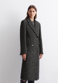 Cappotto doppiopetto in tessuto a pois bianco e nero, di lunghezza media con rever a punta, grandi bottoni e tasche laterali.