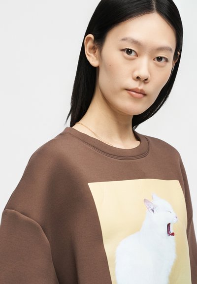 Νέα γυναίκα με ίσια μαύρα μαλλιά φοράει καφέ φούτερ με μεγάλο σχέδιο μιας χασμουρηστής λευκής γάτας σε ανοιχτό κίτρινο φόντο.