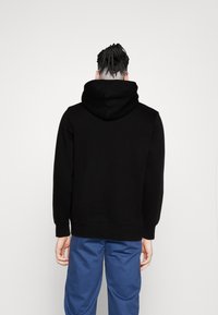 Svart hettegenser laget av myk stoff, med en avslappet passform og ribbestrikkede mansjetter. Personen har på seg blå bukser.