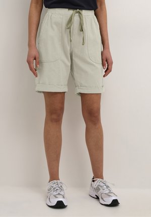 Personne portant un short beige avec cordon et revers roulés ainsi que des chaussures de sport blanches, debout devant un fond clair uni.