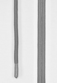 Nära bild av ett vitt tyg med två vertikala grå skosnören och detaljerad sömnad längs kanterna.