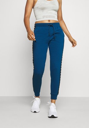 Mujer con pantalones jogger azules con rayas negras en los lados, una camiseta sin mangas corta blanca de Puma y zapatillas blancas, posando frente a un fondo liso.