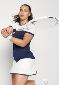 Ναυτικό και λευκό κοντομάνικο μπλουζάκι τένις με στρογγυλή λαιμόκοψη, συνδυασμένο με λευκή φούστα. Ο παίκτης κρατά ένα ρακέτα τένις, έτοιμος να χτυπήσει.