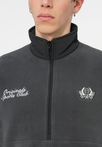 Sivi flis pulover z visokim ovratnikom, polovičnim zadrgo, izvezenim logotipom "Originals Sports Club" in izvezenim grafičnim emblemom na prsih.