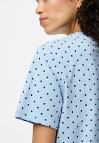 Camiseta de manga corta de color azul claro con lunares negros, con un escote redondo y un corte holgado. Detalle de costura visible cerca del dobladillo.