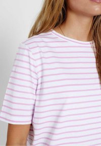 Žena má na sobě bílé tričko s krátkými rukávy a světle růžovými vodorovnými pruhy, odkryté pravé rameno a krk.