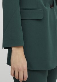 Mörkgrön skräddarsydd blazer med slagkrage, en knappslutning och en ficka med klaff. Slätt, texturerat tyg med en strukturerad passform.