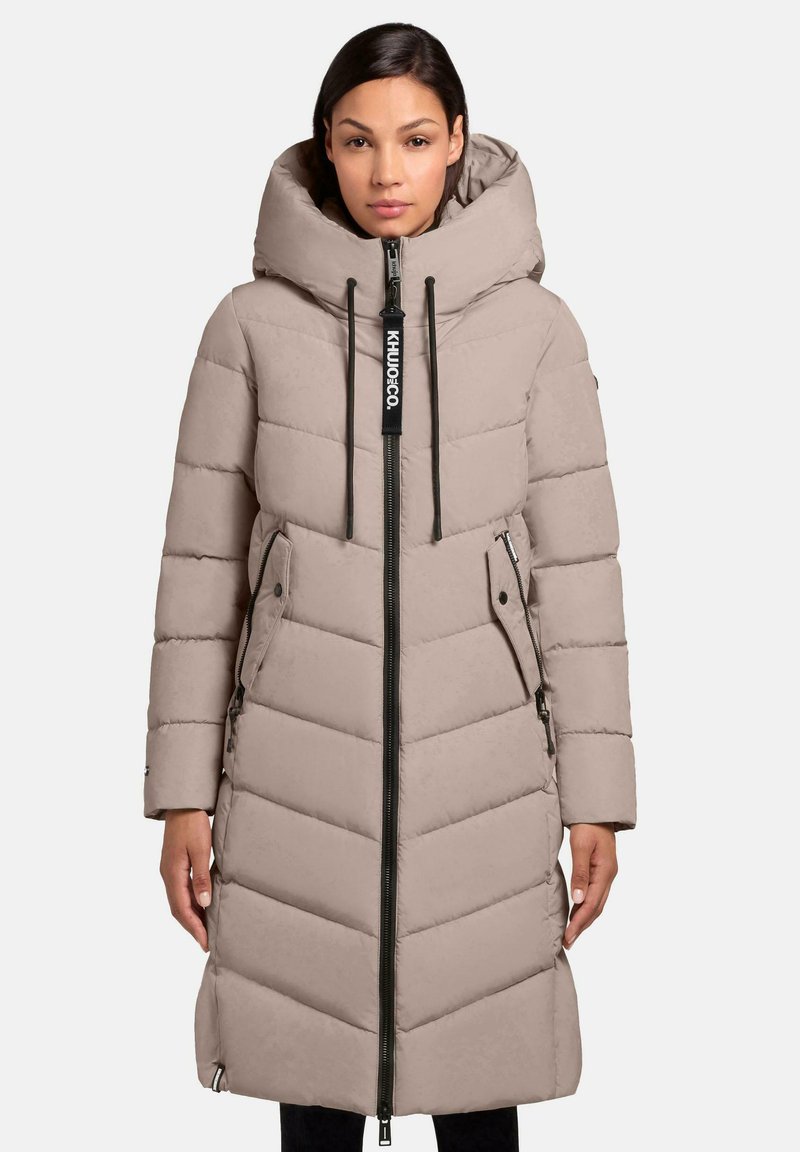 Beige pufferjakke med høj krave, lynlås foran og to sidelommer med lynlås. Har chevron-quilting og justerbare snore i huen.