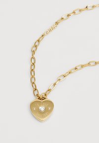 Gouden ketting met een hartvormige hanger waarin "IN LOVE SINCE 1981" is gegraveerd, en versierd met een centrale strasssteen.