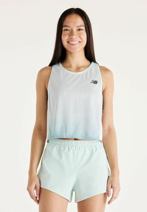 Femme souriante portant un débardeur de sport bleu clair et un short assorti, debout devant un fond blanc, les mains relâchées sur les côtés.