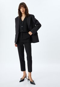 Zwarte streeppakken met een lange blazer met brede revers, een aansluitende vest met twee knopen en taps toelopende broeken, aangevuld met spitse zwarte hakken.