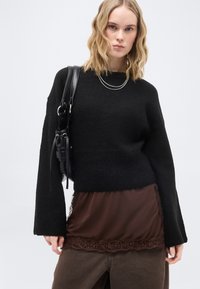 Ξανθιά γυναίκα με μαύρο πουλόβερ, καφέ μπλούζα με δαντέλα και σκούρο παντελόνι, κρατώντας μαύρη δερμάτινη τσάντα ώμου σε λευκό φόντο.