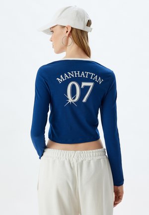 Sininen pitkähihainen crop-top, jossa valkoinen kaulus, takana metallinhohtoisella hopealla painettuna "MANHATTAN 07". Yhdistetty kermaisiin collegehousuihin.