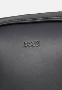 Sac en cuir noir avec logo "BOSS" en relief. Texture lisse avec un subtil détail de couture le long des bords. Design compact et minimaliste.