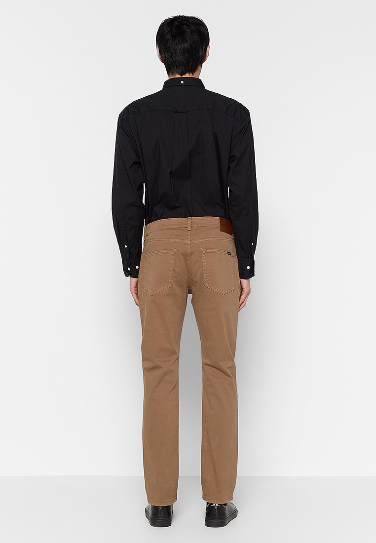 Chino en coton marron avec une coupe droite, arborant un patch en cuir sur la taille et les poches arrière. Porté avec une chemise noire à manches longues.