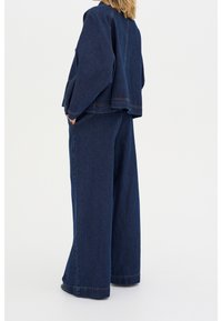 Kvinna i en lös passande mörkblå denimjacka och vida jeans, stående med händerna i fickorna mot en enfärgad bakgrund.