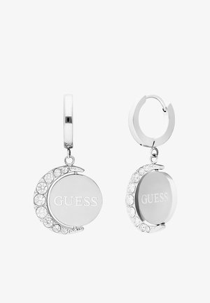 Boucles d'oreilles en argent en forme de créoles avec un pendentif rond portant l'inscription "GUESS", agrémentées de cristaux transparents sur un côté.