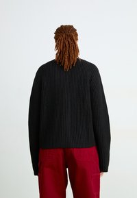 Schwarzer gerippter Strickpullover mit lockerer Passform, langen Ärmeln und rundem Ausschnitt, kombiniert mit roten Cargo-Hosen.