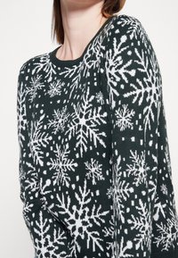 Pullover verde scuro con motivo a fiocchi di neve bianchi. Collo rotondo, maniche lunghe, texture lavorata a maglia, con diversi disegni di fiocchi di neve distribuiti su tutta la superficie.
