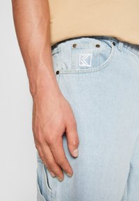 Vaqueros de denim azul claro con bolsillos frontales, remaches de latón y una etiqueta con logotipo blanca. La tela presenta un acabado texturizado. La mano descansa sobre el muslo.