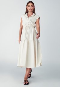 Kreemikas, hõlmik, maksikleit, millel on krae, reguleeritav tõmbluku talje ja kihiline seelik. Kandmisele on lisatud rihmadega mustad sandaalid.