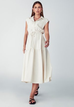 Kremno obarvana, brezrokavna maksi obleka s kragnim ovratnikom, nastavljivim pasom z vrvico in večplastno krilo. Obuta v črne sandale s trakovi.