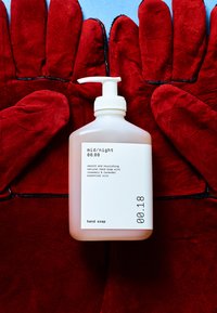 Doorzichtige plastic fles met witte pomp, gevuld met lichtroze vloeibare handzeep met het label "mid/night." Omgeven door rode, getextureerde handschoenen.