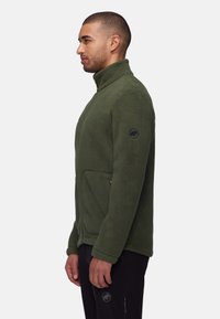 Green fleece jacket with high collar, zip pockets, and a circular logo on the sleeve. Soft texture, casual fit, and solid design.