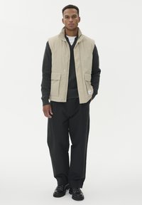 Muškarac nosi bež bezrukavnu jaknu preko tamnosive veste, labave crne hlače i crne čizme, stojeći pored bijele pozadine.