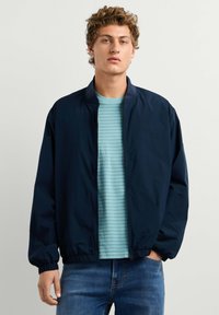 Jeune homme aux cheveux bouclés portant une veste zippée bleu marine sur un t-shirt rayé bleu clair et un jean bleu, debout devant un fond uni.